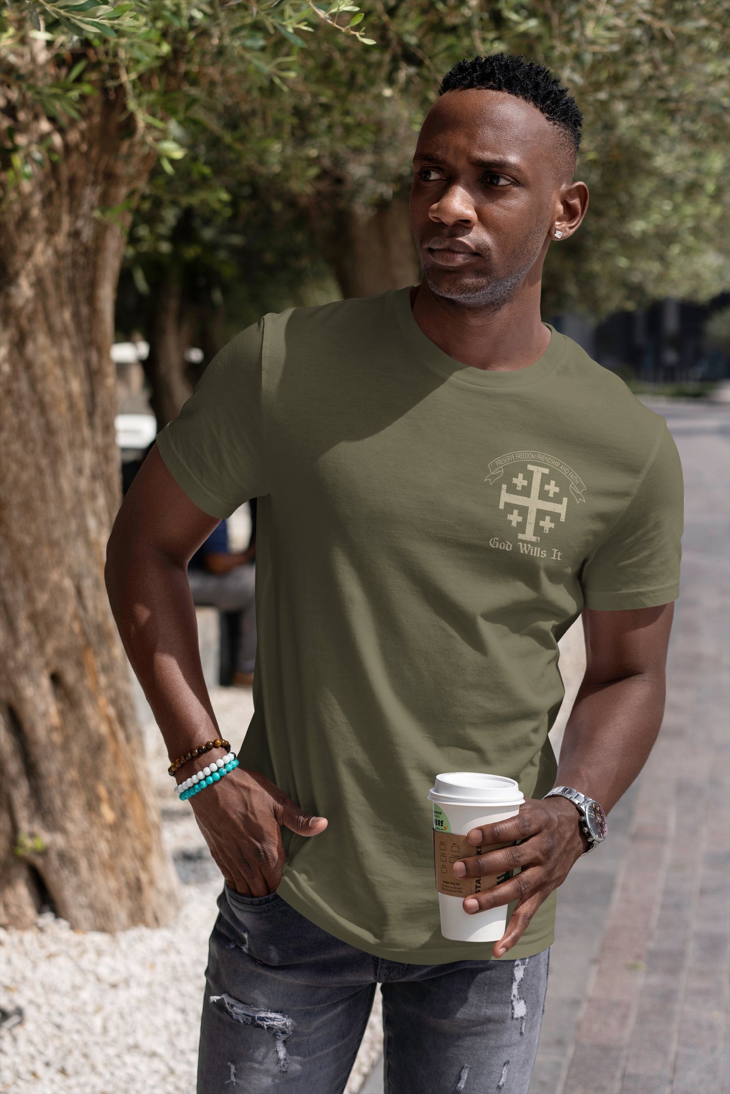 Man holding a coffee cup outdoors standing near a tree in a suburban setting wearing a military green short sleeve t-shirt with a Cross logo on the left chest position with 4 smaller crosses and a banner above the cross with the motto in Latin, "You will preserve freedom, friendship, and faith." Under the cross is printed 'God Wills It' in cream colored print