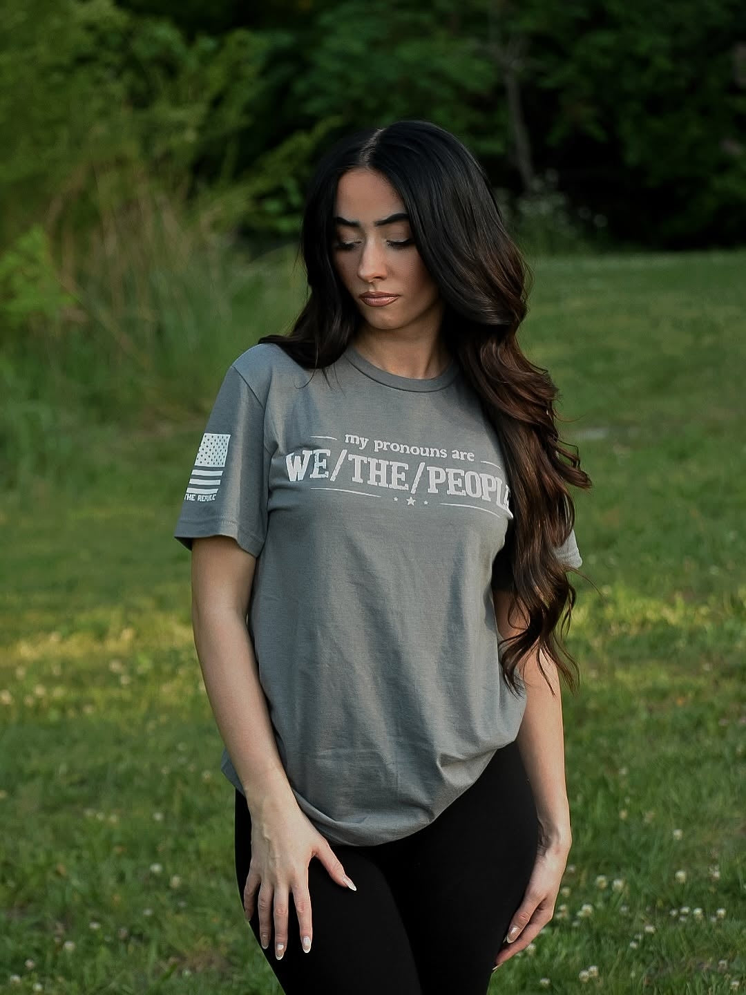 Woman wearing a gray t-shirt with white text stating "my pronouns are we / the / people" in a grassy outdoor setting