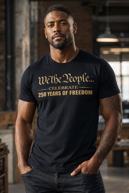 Man wearing a black short sleeve t-shirt with gold text reading 'We The People ... celebrate 250 Years of Freedom' in a rustic indoor setting