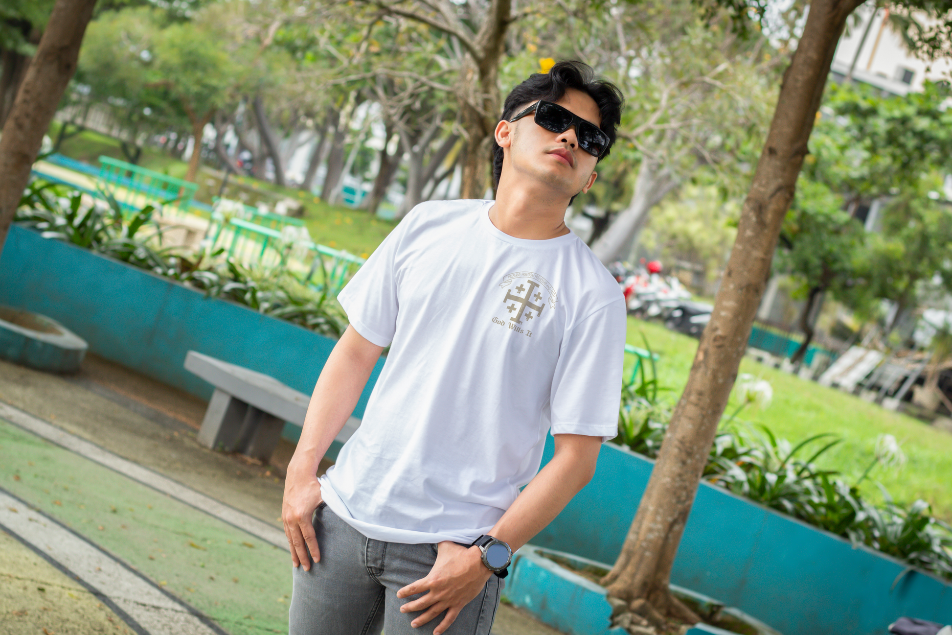 Man wearing sunglasses in park setting with a short sleeve white t-shirt with a Cross logo on the left upper chest position with 4 smaller crosses and a banner above the cross with the motto in Latin, "You will preserve freedom, friendship, and faith."  Under the cross is printed 'God Wills It' in cream colored print