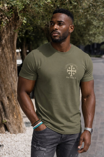 Man standing outside by a tree wearing a military green short sleeve t-shirt with a Cross logo on the left chest position with 4 smaller crosses and a banner above the cross with the motto in Latin, "You will preserve freedom, friendship, and faith."  Under the cross is printed 'God Wills It' in cream colored print