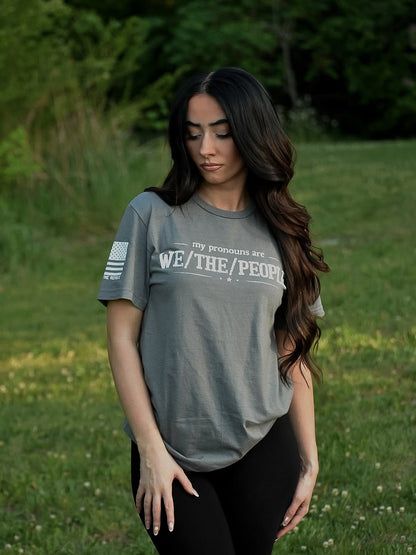 Woman wearing a gray t-shirt with white text stating "my pronouns are we / the / people"  in a grassy outdoor setting
